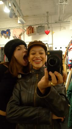 two women taking a selfie in a clothing store with one holding a camera and the other looking at her phone