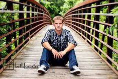 a young man is sitting on a bridge in the woods and posing for a photo