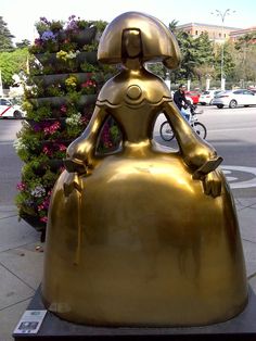 a gold statue sitting on top of a metal base next to flowers and buildings in the background