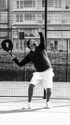 black and white photograph of a man playing tennis on a court with buildings in the background