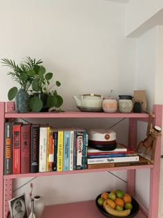 a pink shelf with books and bowls of fruit sitting on it's sides next to a potted plant