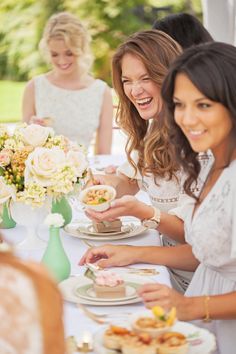 three women sitting at a table with plates of food and flowers in vases on the table