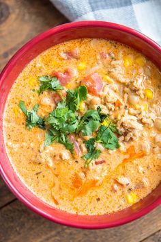 a red bowl filled with soup and garnished with cilantro, corn, and parsley