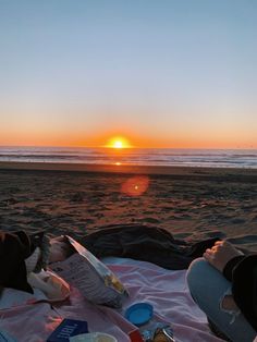 the sun is setting at the beach with people laying in bed and eating food on the sand