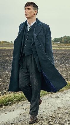 a man walking down a dirt road wearing a long coat and tie with his hands in his pockets