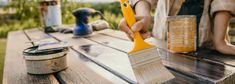 a person with a yellow paintbrush on top of a wooden table next to cans