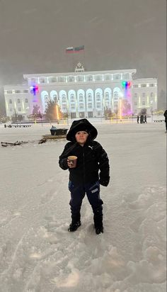 a young boy standing in the snow with a cup of coffee and building in the background