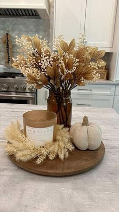 a wooden plate topped with a vase filled with dried flowers and leaves on top of a kitchen counter