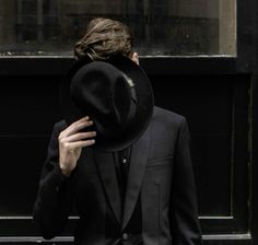 a man in a black suit and hat walks down the street with his hand on his head