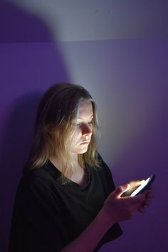 a woman holding a cell phone in her hand and looking at the screen while standing against a purple wall