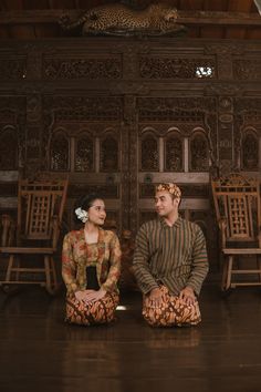 a man and woman sitting on the floor in front of wooden chairs with carved walls behind them