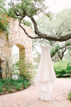 a wedding dress hanging from a tree in front of a stone wall and brick walkway