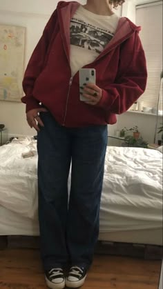 a woman standing in front of a bed wearing a red jacket and blue jeans, holding a cell phone