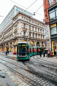 a green train is on the tracks in front of a building and people are walking around