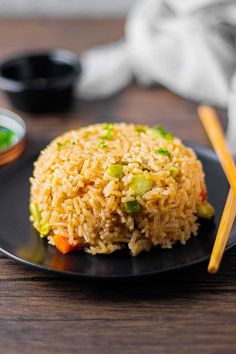 a black plate topped with rice and veggies next to chopsticks on a wooden table