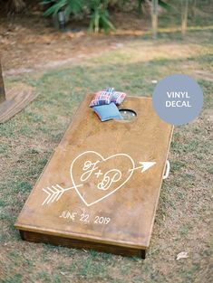 a wooden cornhole board with an arrow and heart drawn on it, sitting in the grass