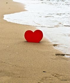 a red heart sitting on top of a sandy beach next to the ocean's edge