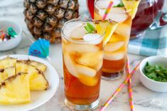 two glasses filled with drinks next to pineapple slices and other fruit on the table