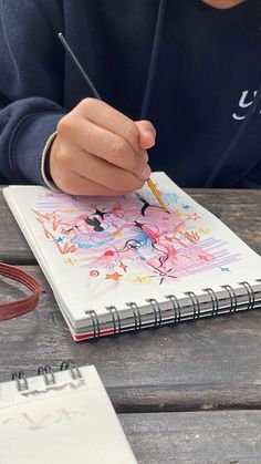 a person sitting at a wooden table with a notebook and pencil in their hand while drawing