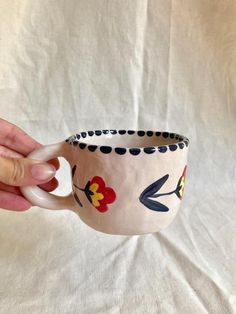 a hand holding a coffee cup decorated with flowers and dots on white fabric, in front of a plain background