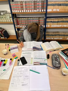 a woman sitting at a desk with books and papers on it, writing in a notebook