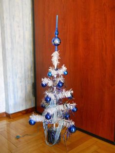 a white christmas tree with blue ornaments on it in front of a wooden wall and floor