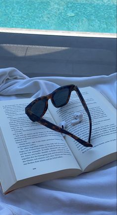 an open book with sunglasses and earbuds laying on it next to a swimming pool