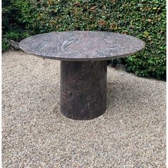 a round table sitting on top of gravel next to a bush filled with green plants