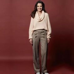 a woman standing in front of a red background wearing brown pants and a beige sweater