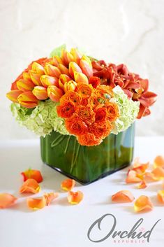 an arrangement of orange and green flowers in a square vase with petals scattered around it