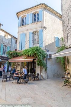 people are sitting at tables outside in front of an old building with blue shutters