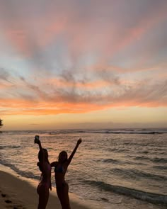 two women are standing on the beach with their arms in the air as the sun sets