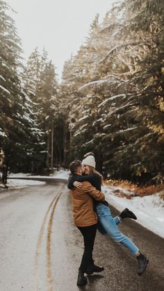 two people are hugging on the road in front of snow covered trees and evergreens