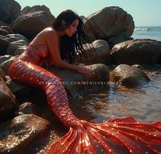 a woman sitting on top of rocks next to the ocean wearing a red mermaid tail