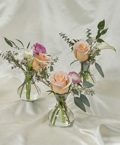 three vases filled with different types of flowers on a white cloth covered tablecloth