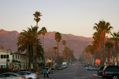 palm trees line the street in front of mountains and buildings at sunset with people walking on the sidewalk