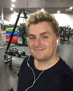 a man with headphones on taking a selfie in a gym room full of exercise equipment