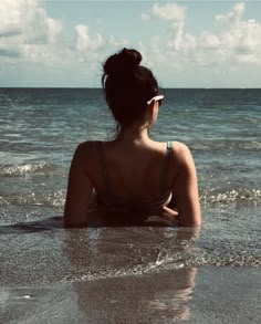 a woman sitting in the water at the beach looking out into the ocean with her back to the camera