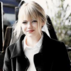 a woman with blonde hair wearing a black jacket and white shirt is standing in front of a car