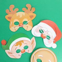 three paper masks with santa hats and reindeer noses on green tableclothed surface next to doughnuts