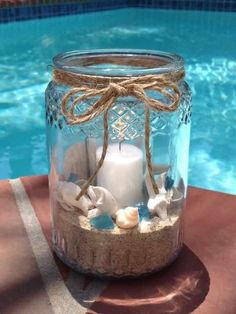 a mason jar filled with sand, shells and a lit candle next to a swimming pool