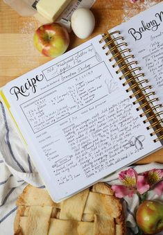 an open recipe book sitting on top of a wooden table next to sliced apples and pies