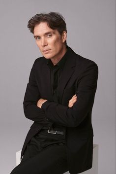 a man in a black suit sitting on a white chair with his arms crossed and looking at the camera