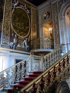 an ornate staircase with red carpet and gold railings in front of a painting on the wall