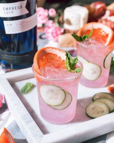 two glasses filled with cucumber and oranges on a tray next to a bottle