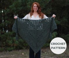 a woman holding up a green crochet shawl in front of some trees