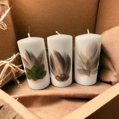 three white candles with feathers on them sitting next to a brown box and some string