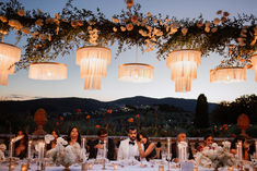 a group of people sitting at a table with candles in front of them and flowers hanging from the ceiling