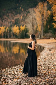 a pregnant woman standing in front of a body of water surrounded by autumn leaves and trees