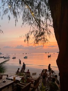 people are sitting at tables on the beach as the sun sets over the water with boats in the distance